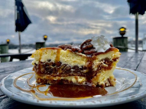 A slice of layered dessert with caramel, nuts, and whipped cream on a plate, set against a cloudy outdoor backdrop.