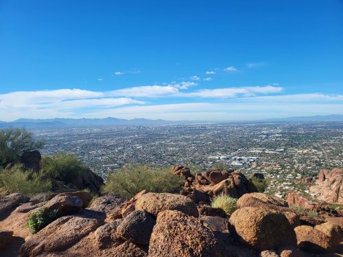 A panoramic view of a cityscape from a rocky mountain, with clear blue skies and distant mountains in the background.