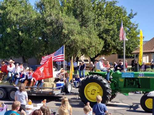 A parade scene with a green tractor, flags, and people on a float, surrounded by spectators and trees.