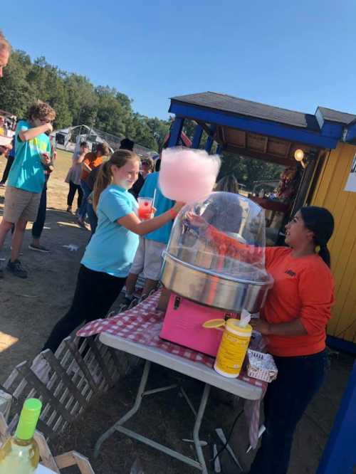 A girl watches as a vendor makes cotton candy at a fair, with people enjoying the event in the background.