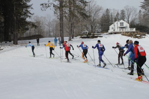 A group of skiers in colorful outfits glides through a snowy landscape, with trees and a house in the background.