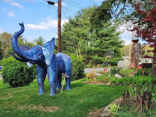 A blue elephant sculpture stands in a garden, surrounded by greenery and a wooden stump in the background.