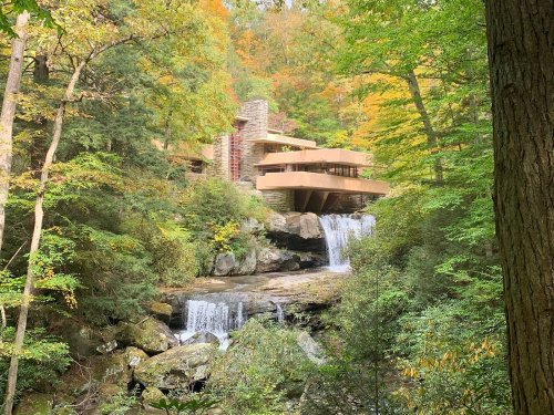 A modern house nestled in a forest, with a waterfall cascading over rocks in the foreground and vibrant autumn foliage.