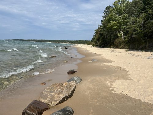 A sandy beach with scattered rocks, gentle waves, and trees lining the shore under a partly cloudy sky.