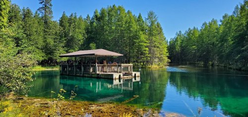 A serene dock on a clear blue lake, surrounded by lush green trees under a bright blue sky.