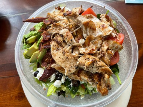 A clear bowl filled with a salad topped with grilled chicken, bacon, avocado, tomatoes, and mixed greens.