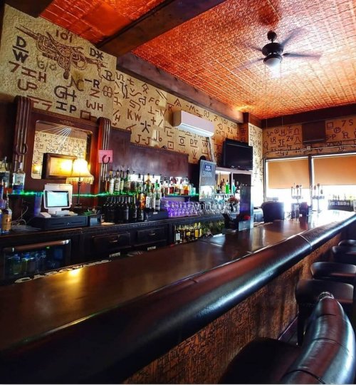 A dimly lit bar with a wooden counter, shelves of liquor, and walls decorated with letters and symbols.