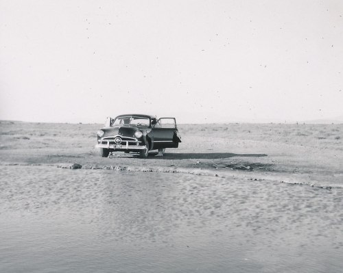 A vintage car parked near a small body of water in a barren landscape, with one door open.