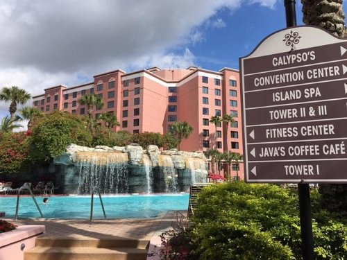 A resort pool with a waterfall, surrounded by palm trees and a sign pointing to various amenities and buildings.