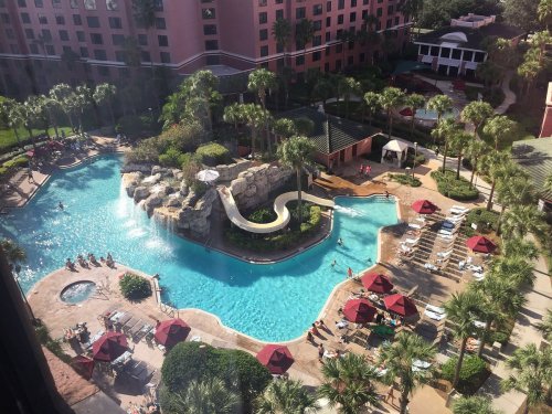 Aerial view of a resort pool with a water slide, surrounded by palm trees and lounge chairs under red umbrellas.