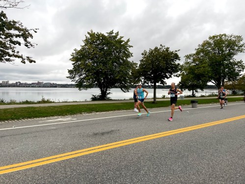 Runners compete along a lakeside path, with trees and a cloudy sky in the background.