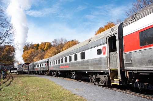 A vintage train with passenger cars parked beside a scenic landscape of autumn trees and a blue sky.