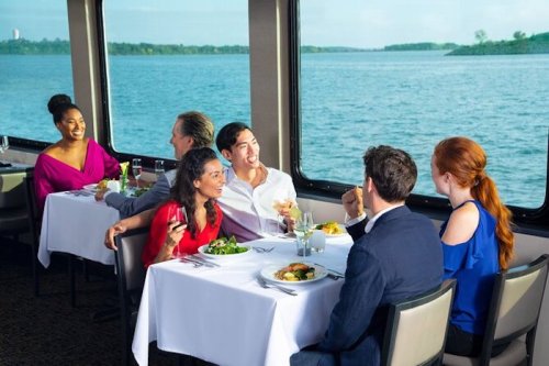 A group of six people enjoying a meal together on a boat, with a scenic view of water and greenery outside.