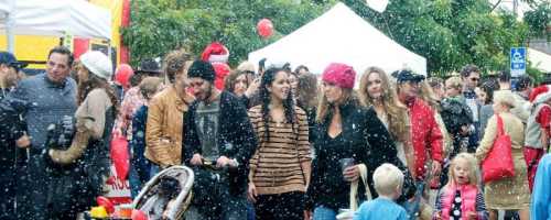 A bustling outdoor event with people walking through light snow, surrounded by tents and festive decorations.