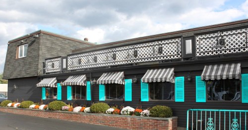 A black building with turquoise shutters and striped awnings, featuring flower beds and a cloudy sky above.