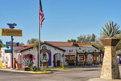 Exterior of El Chaparral restaurant with a flag, palm trees, and a large agave plant in front. Clear blue sky above.