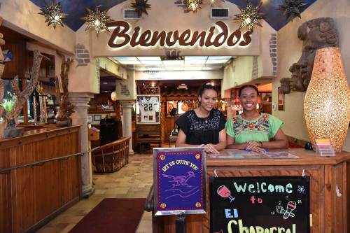 Two women smile behind a reception desk at a vibrant restaurant, with "Bienvenidos" and festive decor in the background.