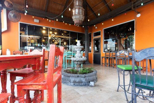 A vibrant restaurant patio featuring colorful wooden furniture, a fountain, and string lights under a wooden ceiling.