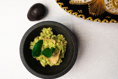A bowl of guacamole topped with green leaves, next to an avocado and a decorative plate.