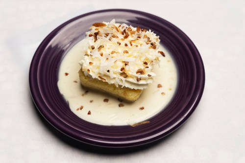 A slice of cake topped with coconut and nuts, served on a purple plate with a creamy sauce.