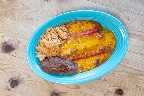 A blue plate with three enchiladas topped with cheese, served with rice and refried beans on a wooden table.