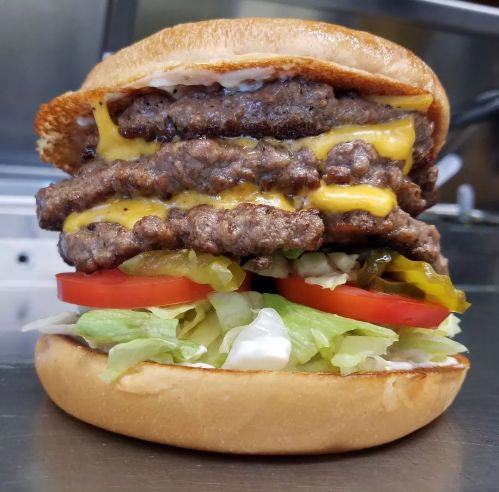 A towering burger with three beef patties, melted cheese, lettuce, tomato, pickles, and mayonnaise on a toasted bun.