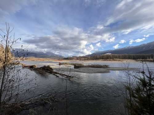 A serene river landscape with mountains in the background, surrounded by trees and a cloudy sky.