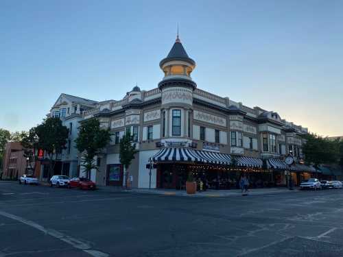 Historic building with a turret, featuring striped awnings and outdoor seating, at a street corner during sunset.