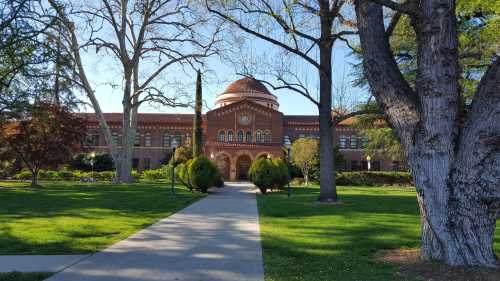 A brick building with a dome, surrounded by trees and greenery, viewed from a paved walkway on a sunny day.