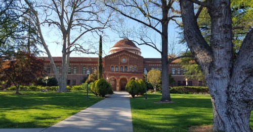 Historic brick building with a dome, surrounded by trees and green lawns, on a sunny day. Pathway leads to the entrance.