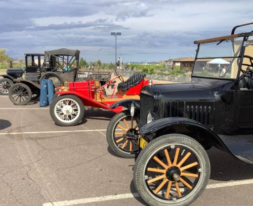 A row of vintage cars parked, including a black car and a red one, against a cloudy sky.