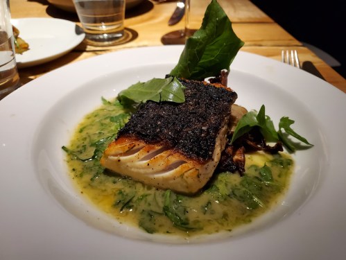 A beautifully plated piece of seared fish on a bed of green sauce, garnished with fresh herbs and greens.