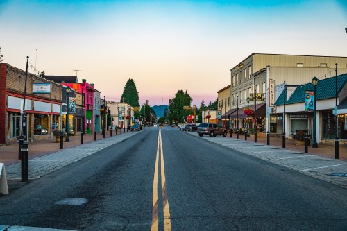 A quiet street in a small town at dusk, lined with shops and trees, with mountains in the background.