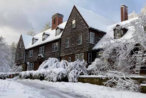 A large, snow-covered house with a steep roof and surrounding trees blanketed in white snow.