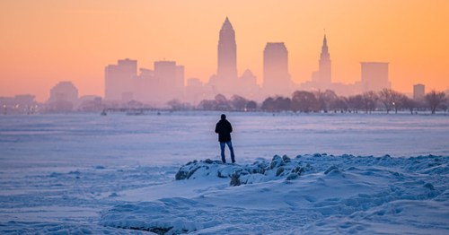 A person stands on a snowy shore, silhouetted against a colorful sunset over a city skyline.