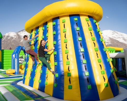Two people climb a colorful inflatable wall at an outdoor amusement park with mountains in the background.