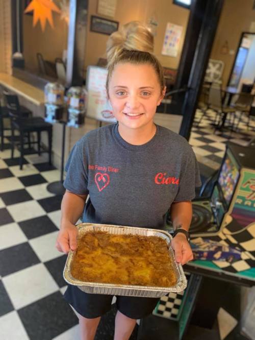 A smiling woman holds a tray of food in a diner with a checkered floor and casual seating in the background.