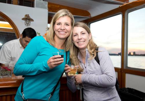 Two smiling women hold drinks on a boat, with a scenic view of the water and sunset in the background.