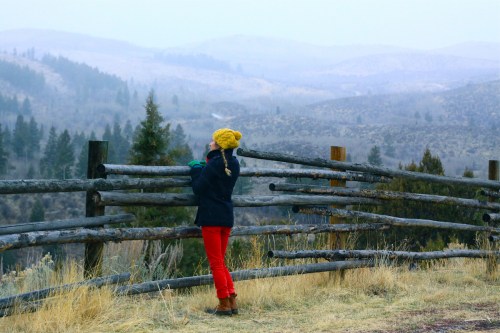 A child in a yellow hat and red pants stands by a wooden fence, gazing at a misty mountain landscape.