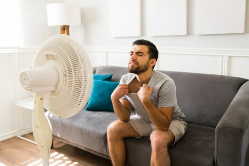 A man sitting on a couch, fanning himself with a shirt, feeling hot in front of a fan.