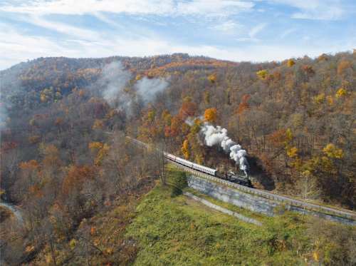 A steam train travels through a colorful autumn landscape with hills and trees, emitting white smoke against a blue sky.