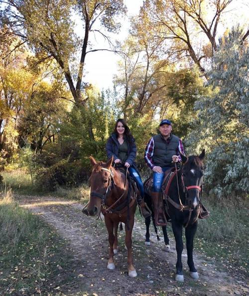 A man and a woman ride horses along a tree-lined path in a scenic outdoor setting.