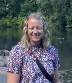 A smiling woman with wavy hair stands by a pond, wearing a floral shirt and a crossbody bag. Green trees are in the background.