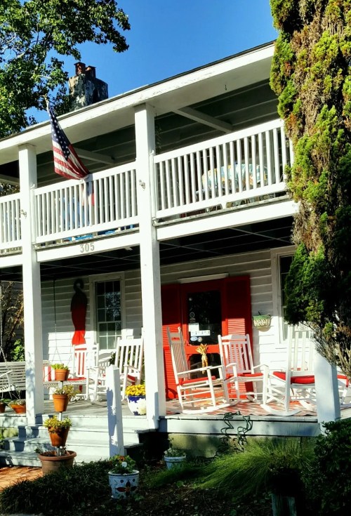 A charming two-story house with a porch, rocking chairs, and an American flag, surrounded by colorful flower pots.