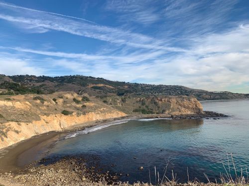 A scenic coastal view featuring cliffs, a sandy beach, and calm waters under a blue sky with wispy clouds.