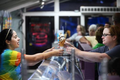 A server hands a large ice cream cone to a customer at an ice cream shop, with people waiting in the background.
