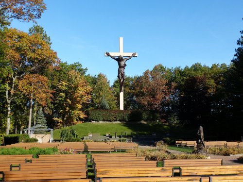 A large crucifix stands on a hill surrounded by trees, with wooden benches arranged in front for gatherings.