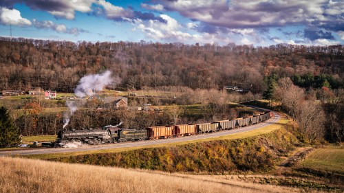A steam locomotive pulls a long train of freight cars through a scenic, wooded landscape under a cloudy sky.