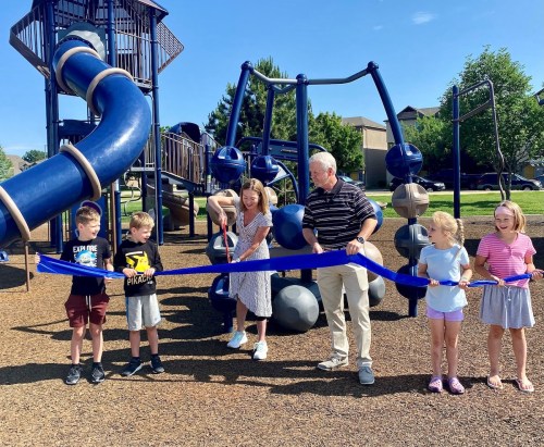 A group of children and adults cut a blue ribbon at a playground opening, with play structures in the background.