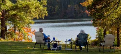 Two people sit in chairs by a lake, surrounded by trees, enjoying a peaceful view on a sunny day.